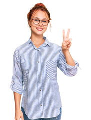 Wall Mural - Young redhead woman wearing casual clothes and glasses smiling with happy face winking at the camera doing victory sign. number two.