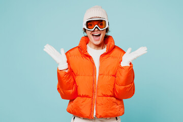 Young excited overjoyed woman wear warm padded windbreaker jacket hat ski goggles mask spread hands look camera travel rest spend weekend winter season in mountains isolated on plain blue background.