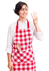 Wall Mural - Beautiful young woman with short hair wearing professional cook apron showing and pointing up with fingers number three while smiling confident and happy.