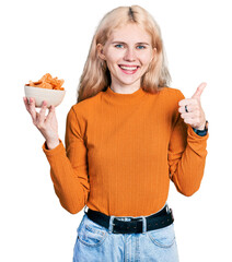 Canvas Print - Young caucasian woman holding nachos potato chips smiling happy and positive, thumb up doing excellent and approval sign