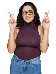 Wall Mural - Brunette young woman wearing casual clothes and glasses gesturing finger crossed smiling with hope and eyes closed. luck and superstitious concept.