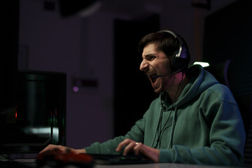 Side view of young male cybersport gamer in green hoodie and headset yelling while playing computer game in dark room