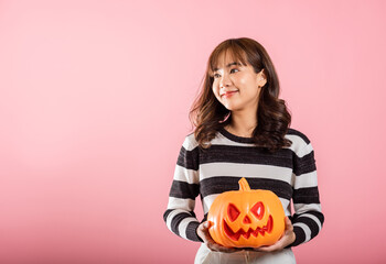 Wall Mural - Portrait of a young Asian woman with a funny Halloween costume, holding model pumpkins, one of which is shaped like a ghost. The studio shot on pink background captures her Halloween enthusiasm.