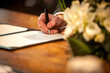 This image captures a close-up of a groom's hand as he signs the wedding register, a pivotal moment symbolizing the legal union of marriage. The warm tones of the wooden table and the soft lighting