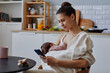 Side view at happy Caucasian mother sitting in kitchen using smartphone while holding napping baby in blanket, copy space