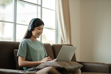 Wall Mural - Young female student wearing headphones sits intently and happily studying online on her laptop on the sofa in the living room at home.