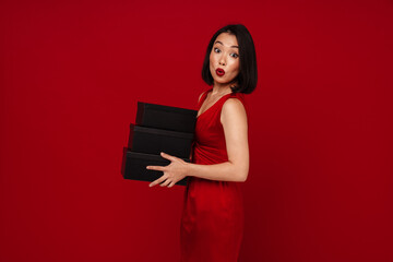 Wall Mural - Surprised woman holding gift boxes and looking at camera isolated over red background