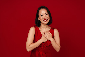 Wall Mural - Lovely asian woman smiling and holding her hands on chest isolated over red background