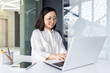 © Liubomir - Successful Asian business woman at workplace inside office, female employee smiling contentedly using laptop at work, woman in light clothes satisfied with achievement results, typing on keyboard.