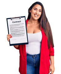 Poster - Young beautiful brunette woman holding clipboard with contract looking positive and happy standing and smiling with a confident smile showing teeth