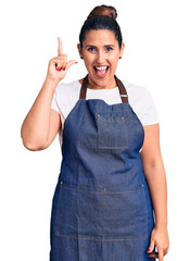 Wall Mural - Young beautiful brunette woman wearing apron pointing finger up with successful idea. exited and happy. number one.