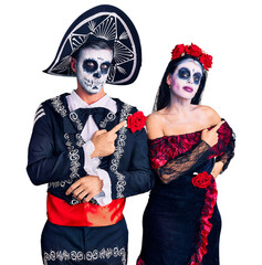 Poster - Young couple wearing mexican day of the dead costume over background pointing with hand finger to the side showing advertisement, serious and calm face