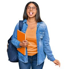 Poster - Young latin girl wearing student backpack and holding books sticking tongue out happy with funny expression. emotion concept.