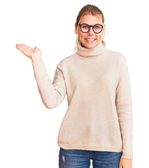 Wall Mural - Young beautiful blonde woman wearing turtleneck sweater and glasses smiling cheerful presenting and pointing with palm of hand looking at the camera.