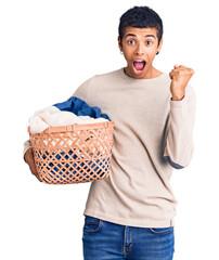 Poster - Young african amercian man holding laundry basket screaming proud, celebrating victory and success very excited with raised arms