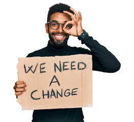 Canvas Print - Young african american man holding we need a change banner smiling happy doing ok sign with hand on eye looking through fingers