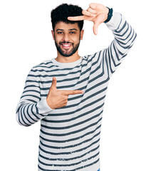 Wall Mural - Young arab man with beard wearing casual striped sweater smiling making frame with hands and fingers with happy face. creativity and photography concept.