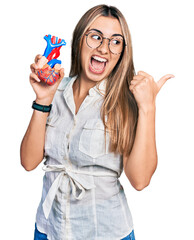 Sticker - Hispanic young woman holding heart organ pointing thumb up to the side smiling happy with open mouth