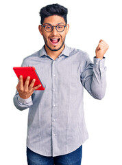 Sticker - Handsome latin american young man holding touchpad screaming proud, celebrating victory and success very excited with raised arms