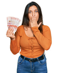 Poster - Beautiful hispanic woman holding 10 colombian pesos banknotes covering mouth with hand, shocked and afraid for mistake. surprised expression