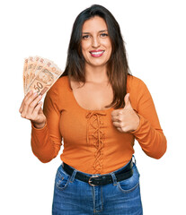 Wall Mural - Beautiful hispanic woman holding 50 brazilian real banknotes smiling happy and positive, thumb up doing excellent and approval sign