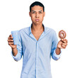 Young hispanic man eating doughnut and drinking coffee puffing cheeks with funny face. mouth inflated with air, catching air.