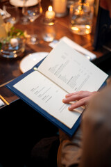 Woman reading a menu in a restaurant. Selective focus on the book. Candles with warm light and dinner set on background