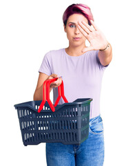 Poster - Young beautiful woman with pink hair holding supermarket shopping basket with open hand doing stop sign with serious and confident expression, defense gesture