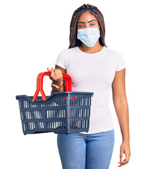 Poster - Young african american woman with braids wearing shopping basket and medical mask looking positive and happy standing and smiling with a confident smile showing teeth