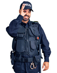 Wall Mural - Young hispanic man wearing police uniform doing ok gesture with hand smiling, eye looking through fingers with happy face.