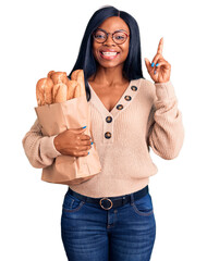 Sticker - Young african american woman holding paper bag with bread surprised with an idea or question pointing finger with happy face, number one