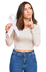 Poster - Young brunette woman holding white ribbon serious face thinking about question with hand on chin, thoughtful about confusing idea