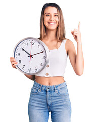 Wall Mural - Young beautiful girl holding big clock surprised with an idea or question pointing finger with happy face, number one