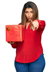 Sticker - Beautiful brunette woman holding gift pointing with finger to the camera and to you, confident gesture looking serious