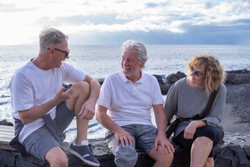 Wall Mural - Group of smiling mature and senior friends sitting back the sea at sunset light, three people talking enjoying freedom and vacation or retirement