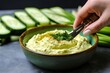 hand using a knife to slice cucumber beside hummus bowl
