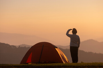 Wall Mural - Traveller is standing by the tent during overnight camping while looking at the beautiful scenic sunset over the mountain for outdoor adventure vacation travel