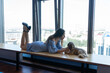 Young woman in blue dress and high heels lies on windowsill looking at dog. Lady rests against window with shih tzu doggy posing for photoshoot