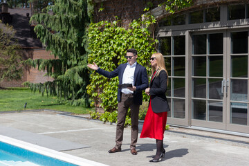 Blonde businesswoman explores available cottage residence from real estate agency. Woman listens to realtor pool description