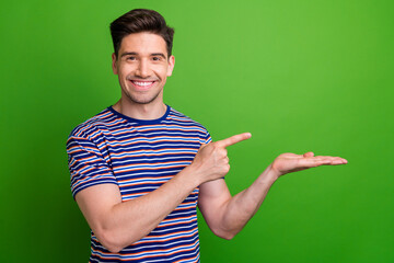 Poster - Photo of positive young man toothy smile indicate finger arm palm hold empty space isolated on green color background