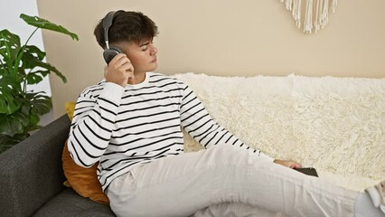 Poster - Cheerful young hispanic man sitting on sofa, relaxed, happily listening to his favorite song at home. enjoying music through smartphone, radiating confidence in his cozy living room.