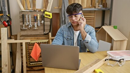 Wall Mural - Attractive young hispanic carpenter engrossed in work, talking on smartphone while accessing computer in his carpentry workshop