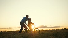 Persistent Boy In Helmet Learns To Ride Bike Under Careful Guidance Of Considerate Daddy. Loving Father Holds Bicycle To Ensure Safety Of Little Son. Family Weekends On Grassy Meadow At Sunset