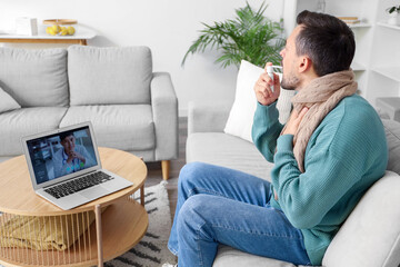 Wall Mural - Ill young man using spray for sore throat while video chatting with doctor on laptop at home