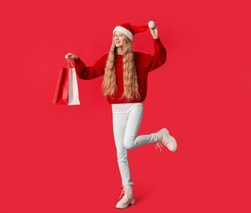 Wall Mural - Young happy woman in Santa hat and warm winter clothes with shopping bags on red background