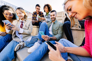 Addicted young people using mobile phones sitting together outdoors. Millennial group of diverse friends looking at cellphone screen, playing video games or enjoying social media content on app.