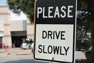 Wall Mural - please drive slowly caption writing text rectangle metal sign with parking lot behind, white and black capital letters with white and black background