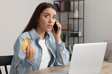 Canvas Print - Confused woman with credit card talking on phone and laptop at table indoors. Be careful - fraud