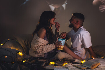 Romantic evening together, calm weekend, slow living. Loving young happy couple having a date at home, watching movies in bed, enjoying each other. Cozy atmosphere, atmospheric lights, leisure