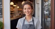 Happy young business woman standing at doorway of her store. Cheerful mature waitress waiting for clients at coffee shop. Successful small business owner in casual wearing grey apron standing